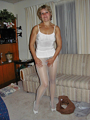 Mature amateur blonde stands confidently in the living room wearing white pantyhose and a white camisole, lifting her skirt to tease her visible bush through the sheer nylon.